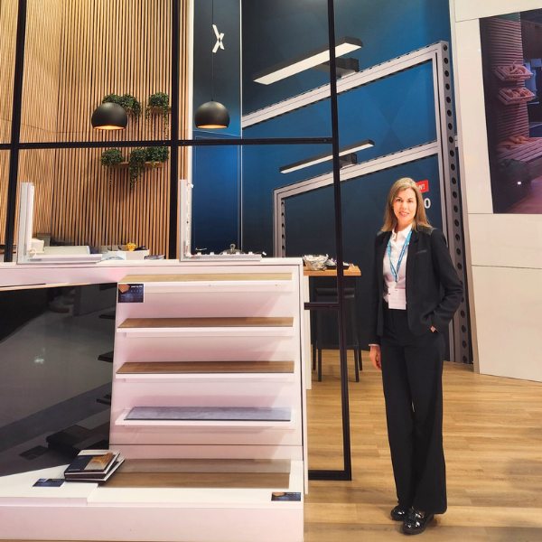 Exhibition booth stand featuring a modern interior design with wooden paneling, hanging black pendant lights, and greenery displayed on the wall. A person in professional business attire stands beside a white product display unit with multiple shelves. The background includes tall blue wall panels with architectural graphics and a high table setup, creating a contemporary and polished exhibition environment