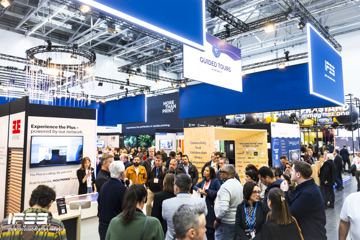 A busy exhibition booth stand at EuroShop featuring branded blue overhead banners, digital displays, and product demonstration areas. A large group of attendees is gathered around a presenter, with various booths and signage visible across the trade show floor, creating a dynamic and crowded exhibition environment